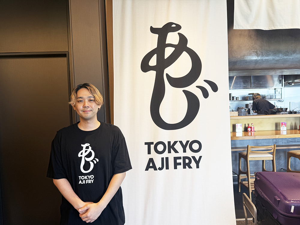 お店のロゴ入りTシャツを着て、店舗の前に笑顔で立つ運営会社の唐橋佑。