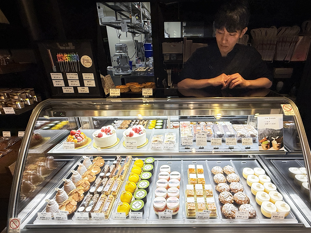 「お菓子と結び」の店内に並ぶケーキのショーケース。モンブランやショートケーキなどの生菓子が美しくディスプレイされ、奥にはパティシエの醍醐憲人さんが立つ様子