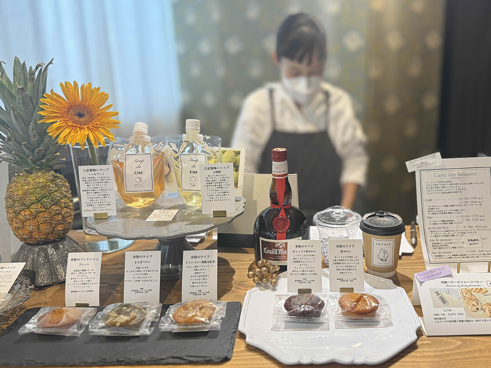 ひまわりやパイナップルが飾られた華やかなカウンターと、その奥に立つARTRIAのオーナー店主・吉崎さんの様子