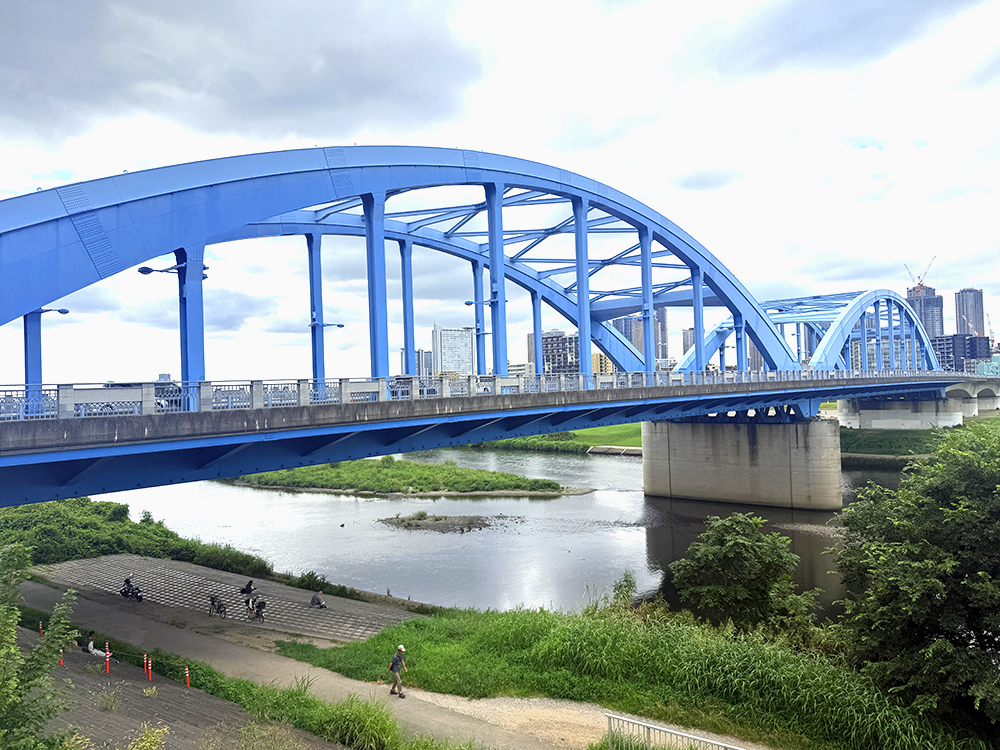 白い雲が広がる空のもと、多摩川に架かる美しく大きな水色のアーチ橋「丸子橋」の全景